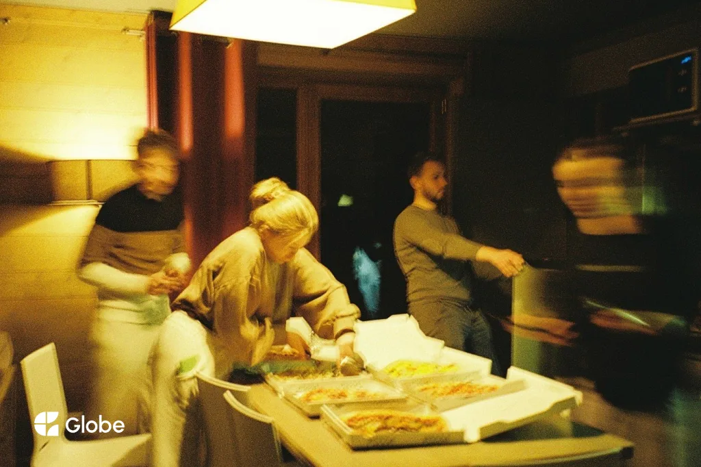 A group of friends gathered around the table of a shared ski apartment at night, helping themselves from several open boxes of pizza under a warm ceiling light