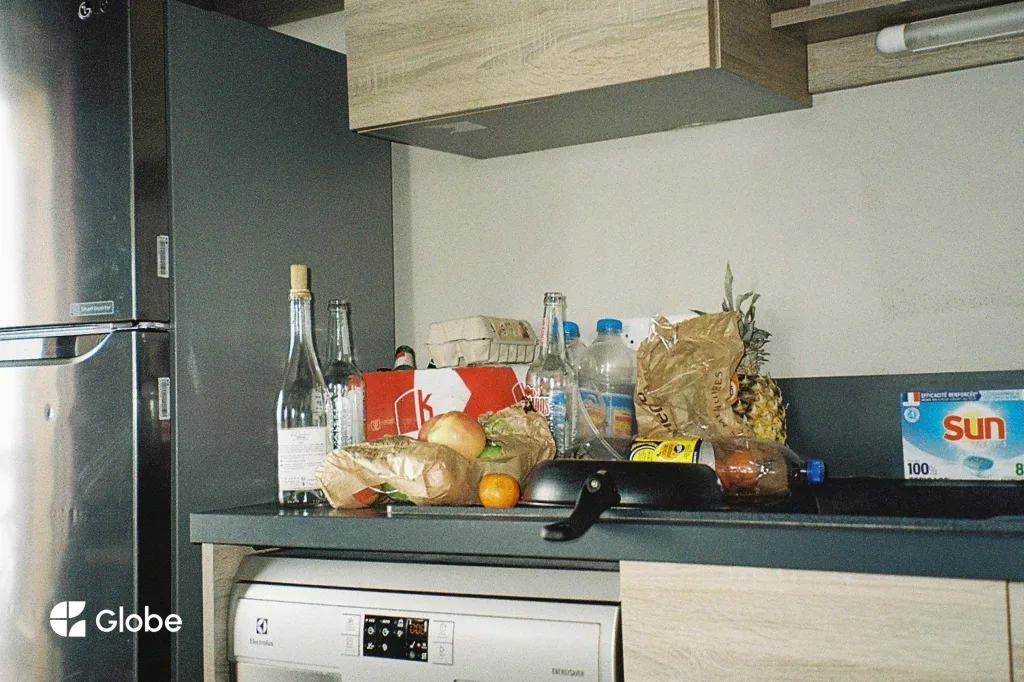 The counter of a self-catering ski apartment kitchen covered with a group supermarket shop: bread in paper bags, wine and beer bottles, large bottles of water, a box of eggs, oranges and a pineapple, and a box of dishwasher tablets