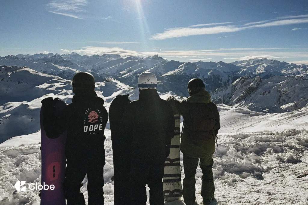 Three snowboarders seen from behind at the top of a ski run, boards standing upright in the snow beside them, looking out over a wide range of snow-covered Alpine peaks under a bright winter sun