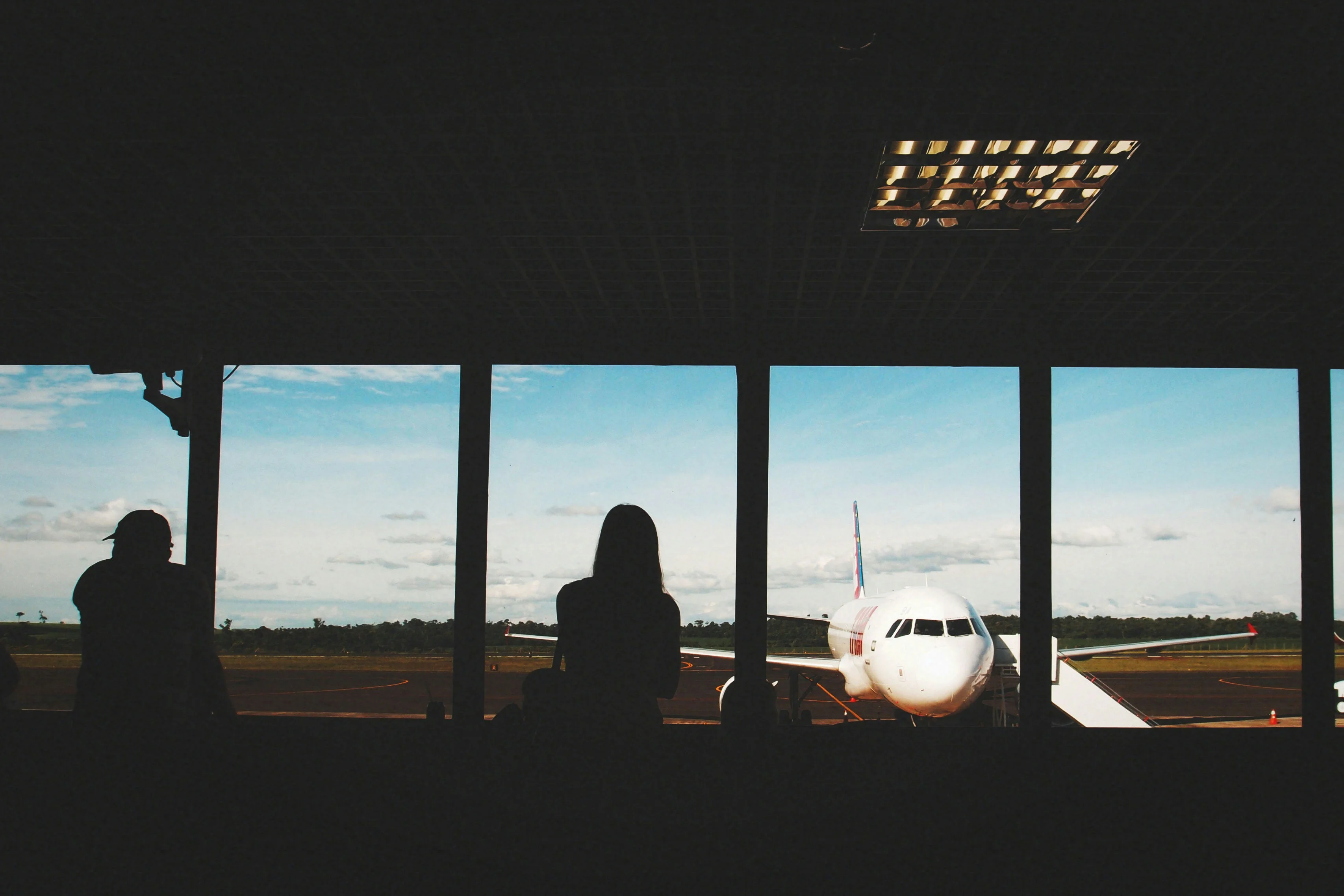 Two friends silhouetted at an airport gate on a group trip, looking out at a plane on the runway
