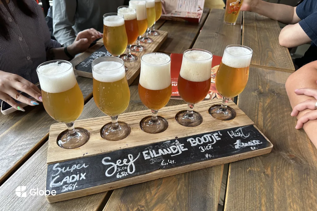 A wooden beer flight board from Antwerpse Brouw Compagnie holding five different Antwerp beers, their names chalked below, shared around a table by a group of friends